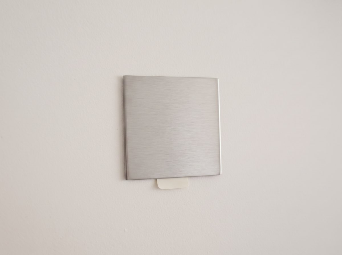The slim metal Facet wall mount stuck to a wall, its removable adhesive pull-tab visible underneath.